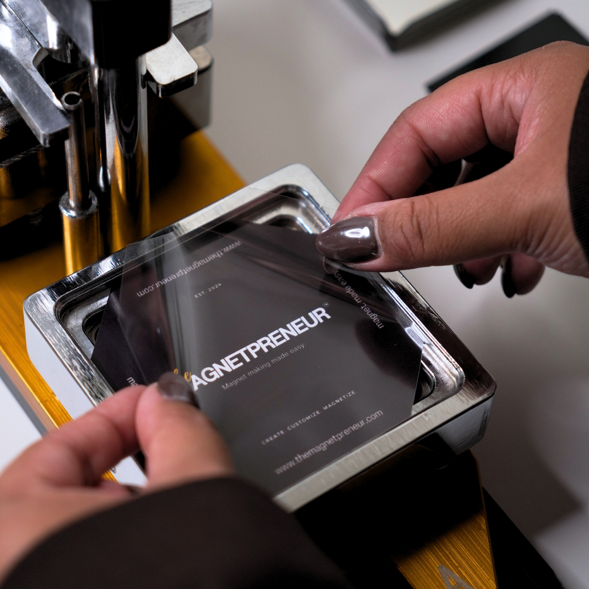 Person using a magnet maker machine to apply a magnet labeled 'The Magnetpreneur' onto a surface.